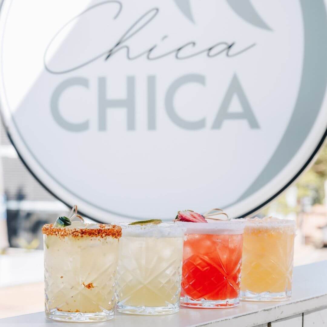 Four cocktail glasses sitting on a bench with a logo in the background that says "Chica Chica"